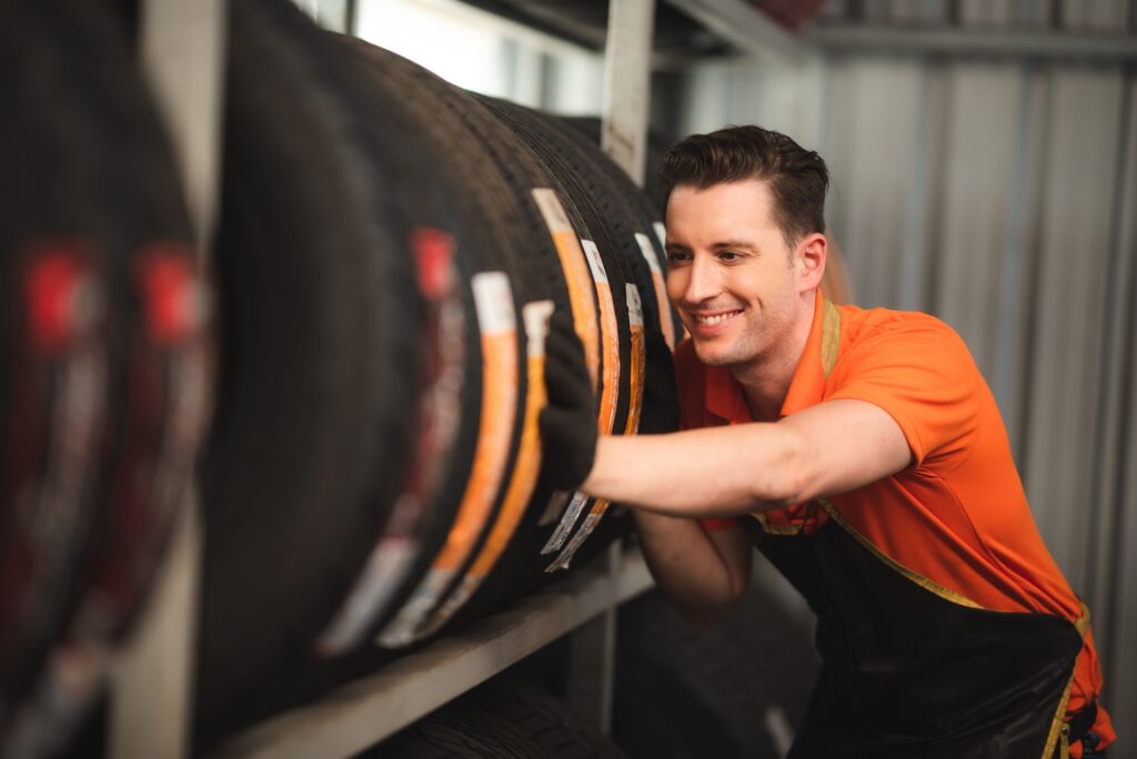 Smiling young car mechanic wearing uniform and hand gloves pushing and checking new tires